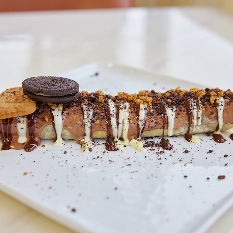 Dessert crepe drizzled with chocolate and white sauces, topped with cookie crumbs and sandwich cookies on a white plate.