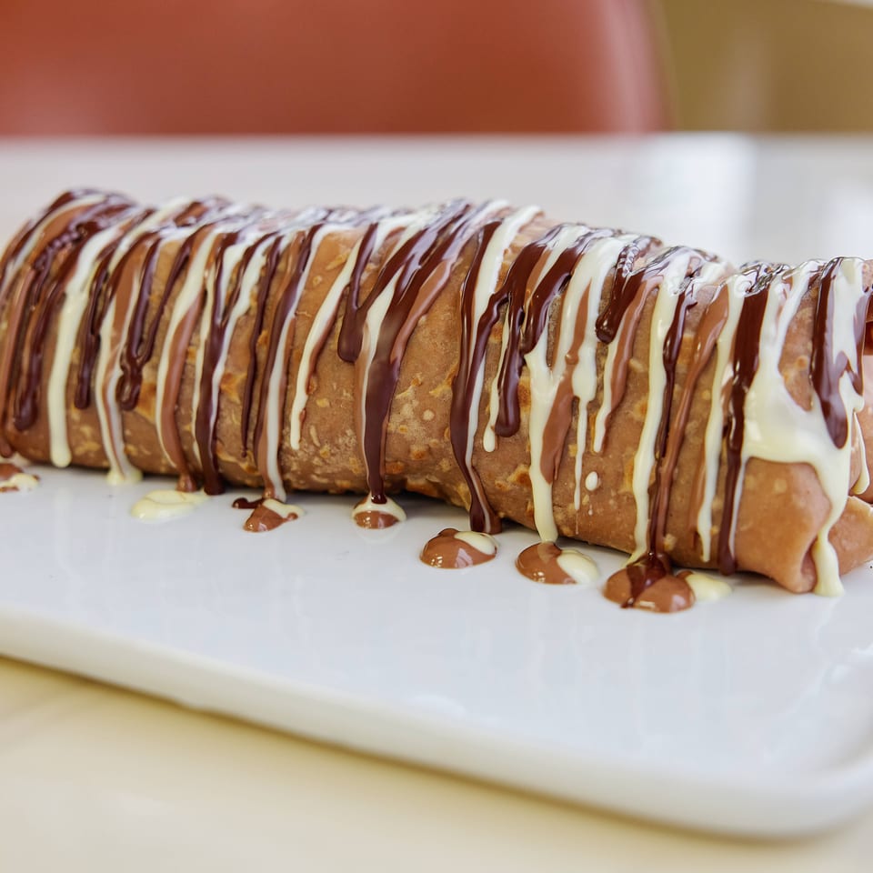 Stacked golden crepes drizzled with chocolate and white icing on a white rectangular plate.