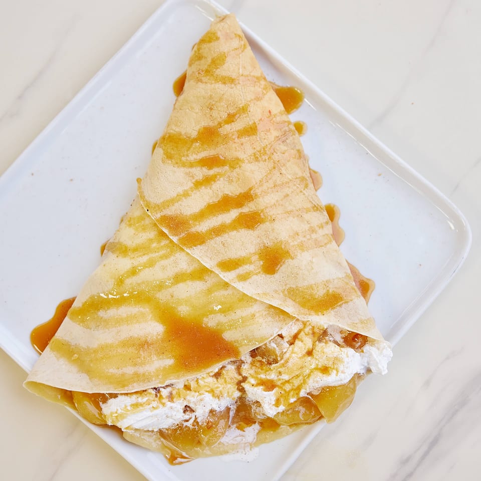Triangular folded crepe drizzled with caramel sauce and filled with whipped cream on a white rectangular plate.