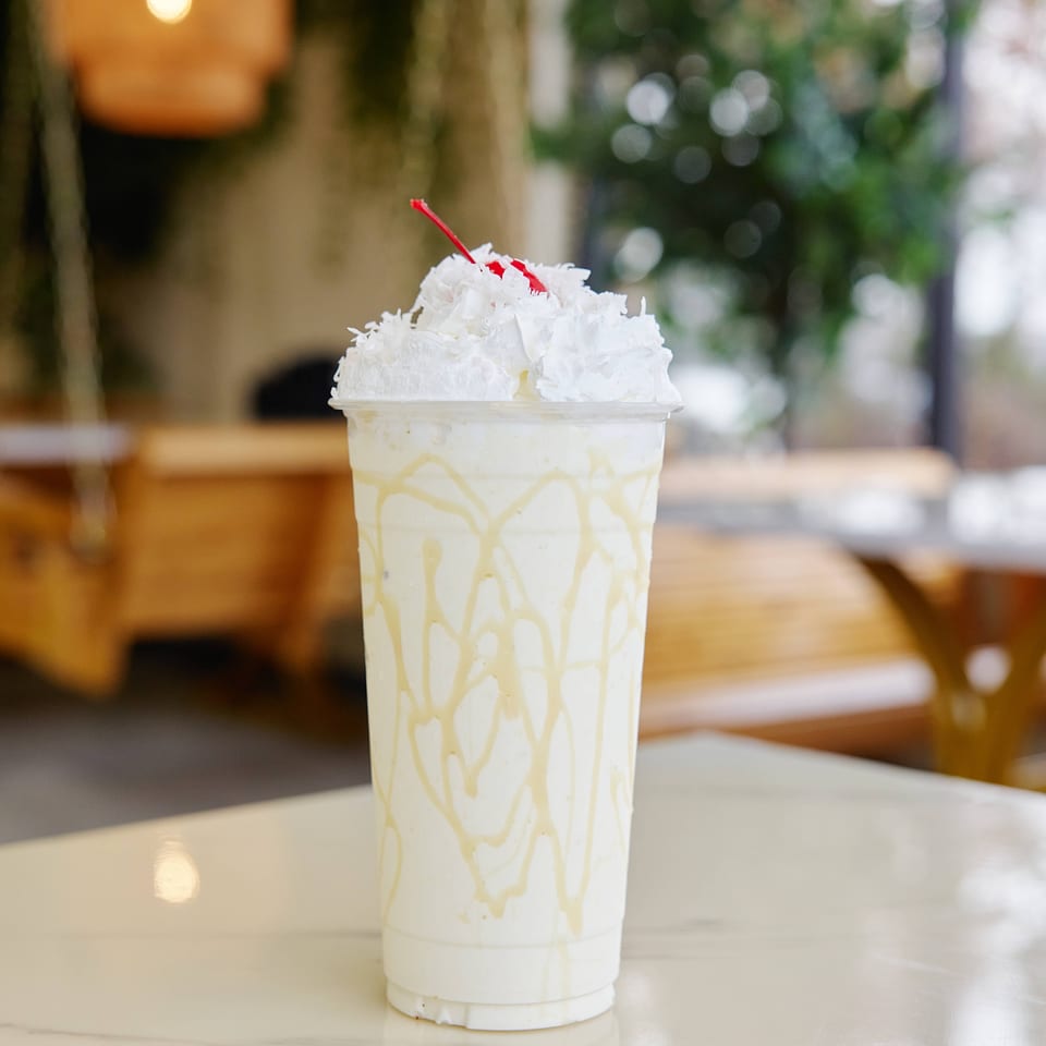 Creamy white dessert drink topped with whipped cream and a red cherry, served in a decorated cup on a café table.