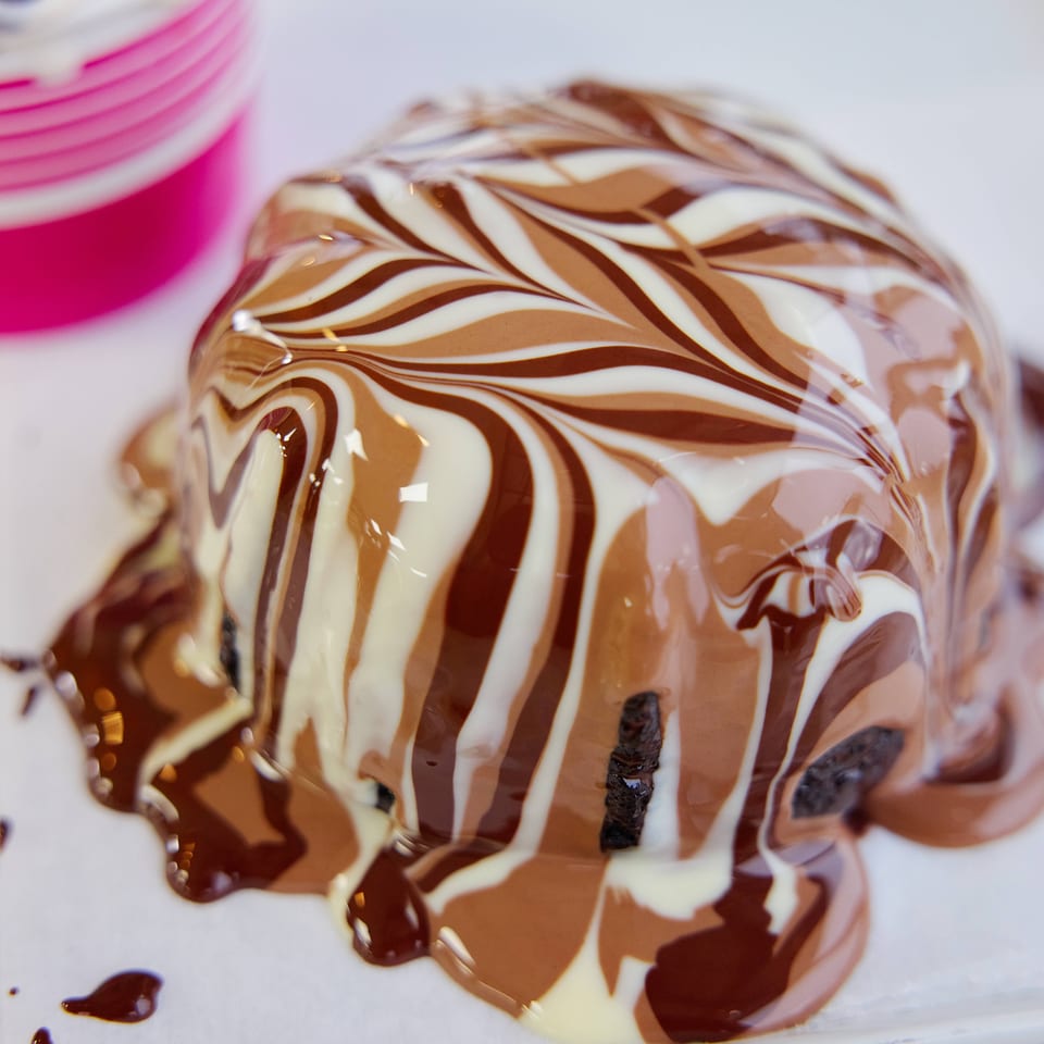 Chocolate dessert dome drizzled with white and dark chocolate, served on a plate beside a pink cup.