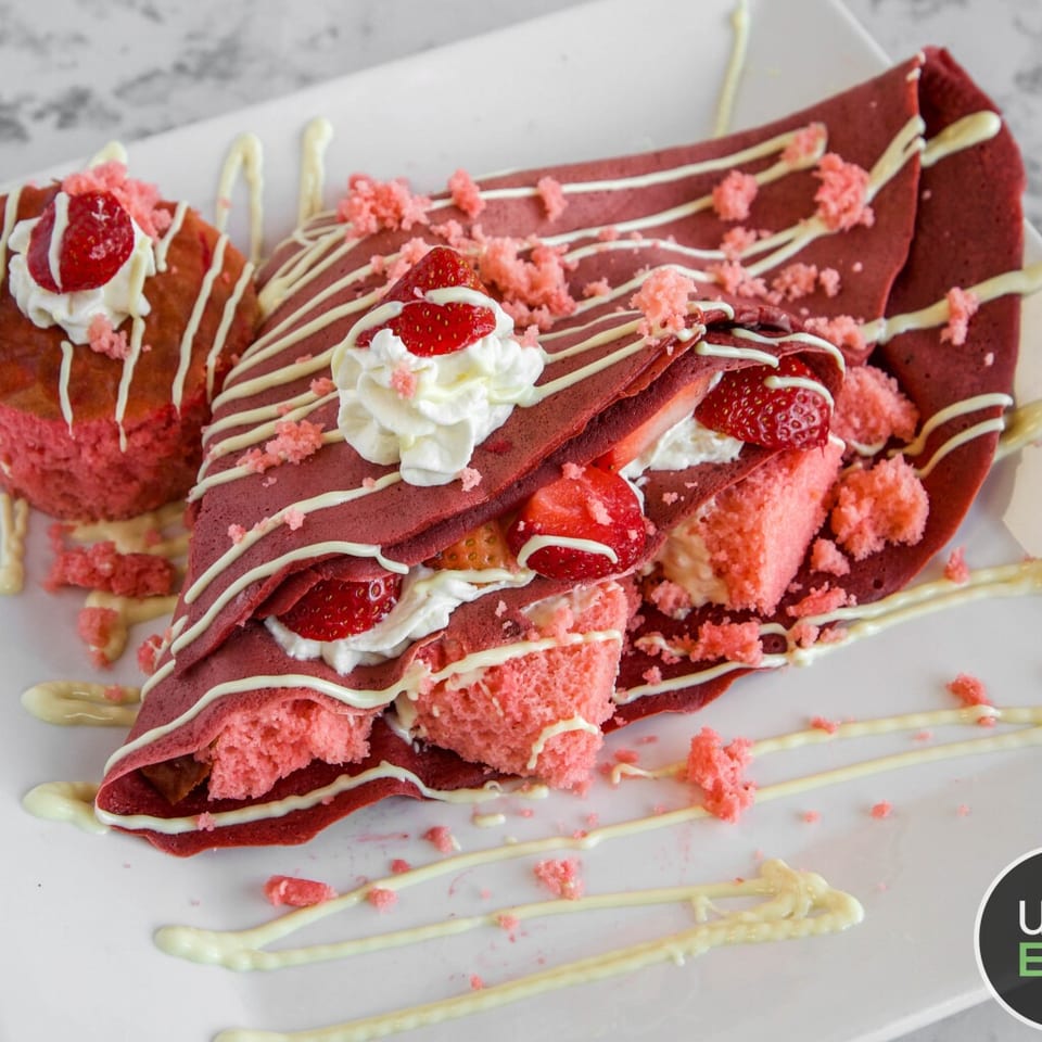 Red velvet crepes layered with strawberries and whipped cream, drizzled with white chocolate and topped with pink crumbs.