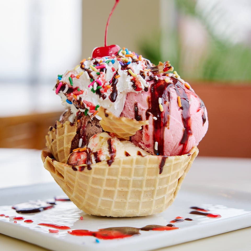 Waffle bowl filled with assorted ice cream, whipped cream, colorful sprinkles, chocolate and strawberry sauces, topped with a cherry.