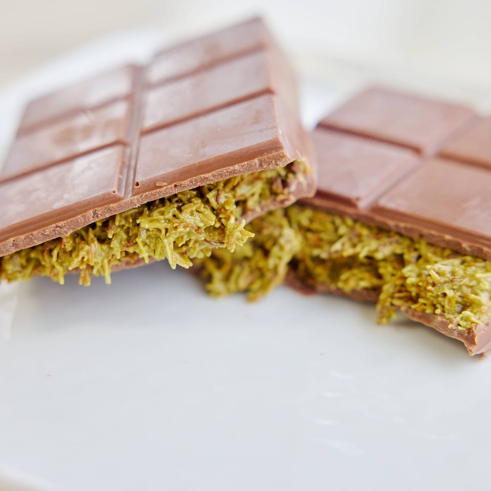 Two milk chocolate bars filled with shredded pistachio paste, one broken open on a white plate.