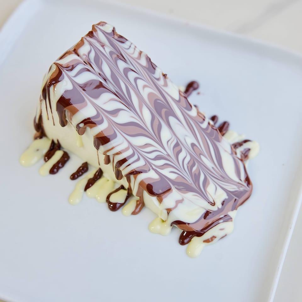 Triangular layered dessert drizzled with chocolate and vanilla sauces, topped with decorative purple and white swirls on a white plate.