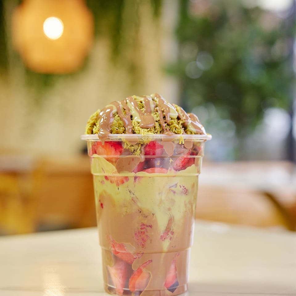 Layered fruit dessert in a clear cup, topped with crushed pistachios and chocolate sauce, on a restaurant table.
