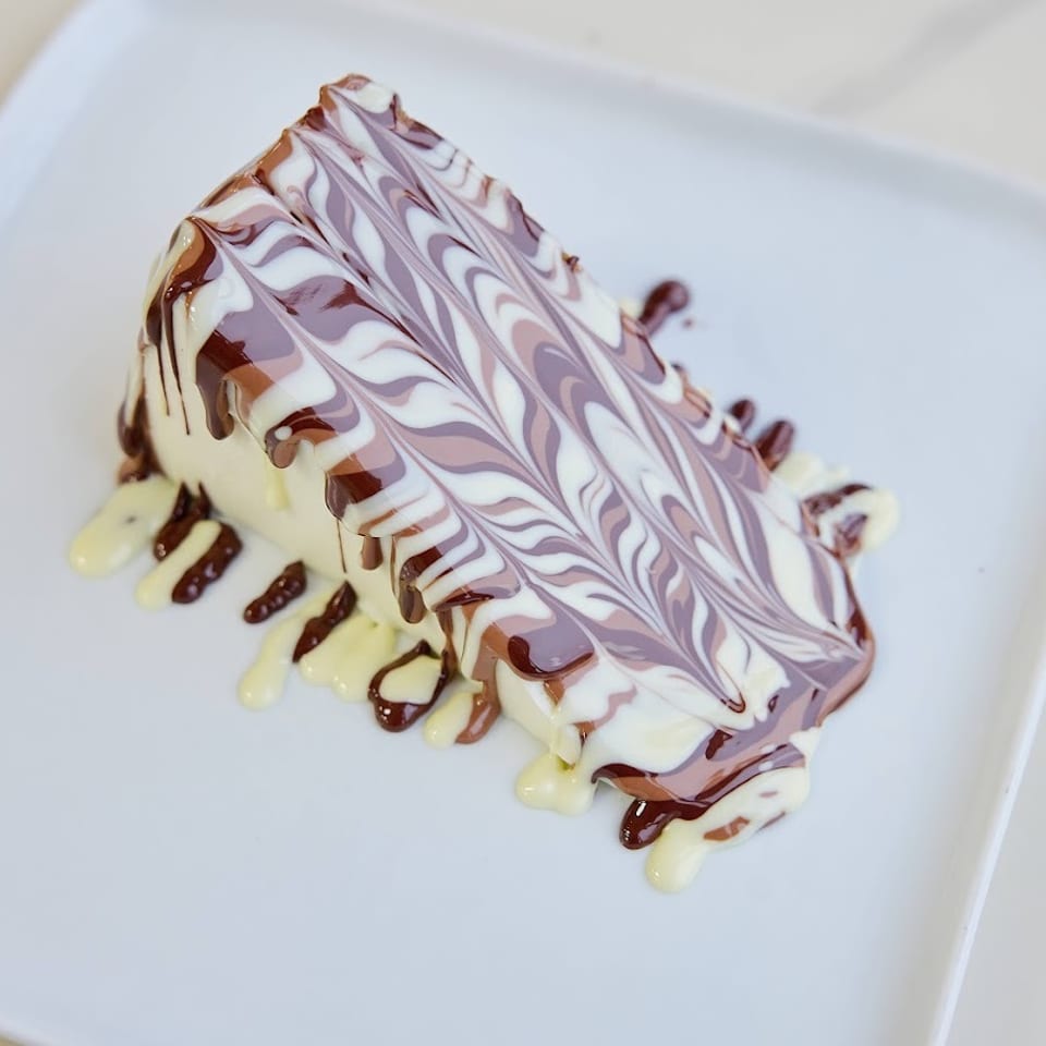 Triangular layered dessert drizzled with chocolate and vanilla sauces, topped with an intricate white and purple swirl pattern.