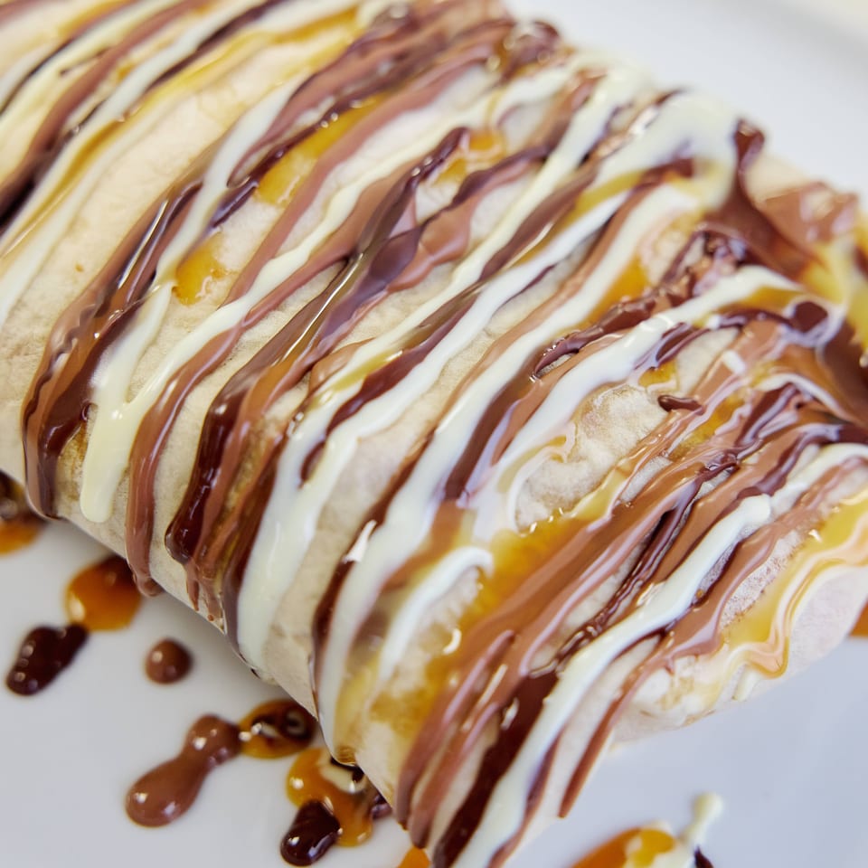 Rolled dessert drizzled with chocolate, vanilla, and caramel sauces on a white plate.