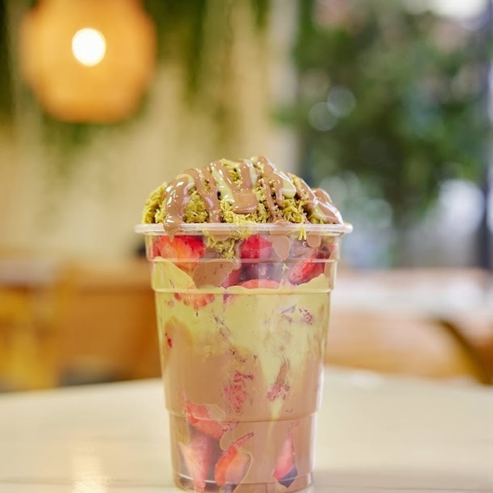 Layered dessert cup with fruit, creamy filling, green pistachio crumbs, and pink sauce on a restaurant table.