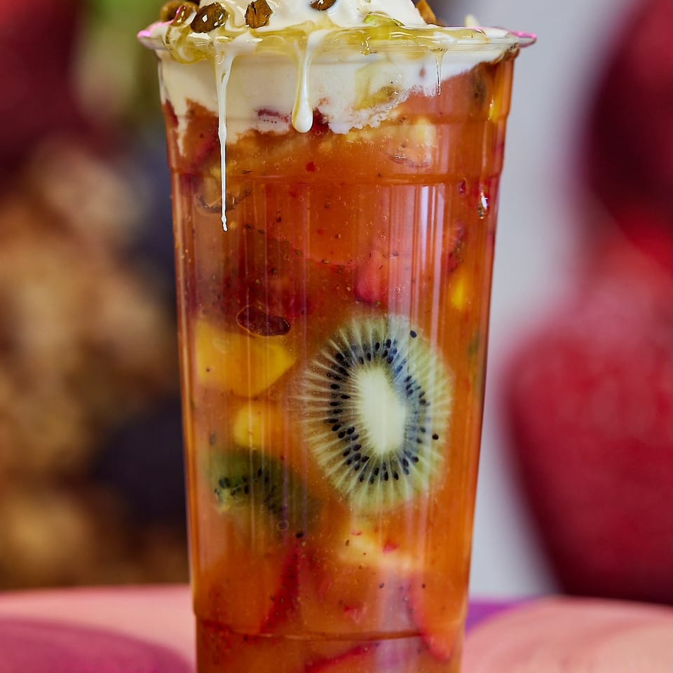 Layered fruit dessert drink with visible kiwi and berries, topped with whipped cream, pistachios, and honey in a glass.