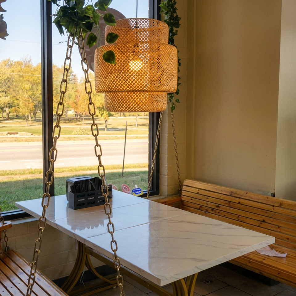 Bright restaurant seating area with swing benches, marble table, hanging wicker lamp, plants, and windows overlooking a park.