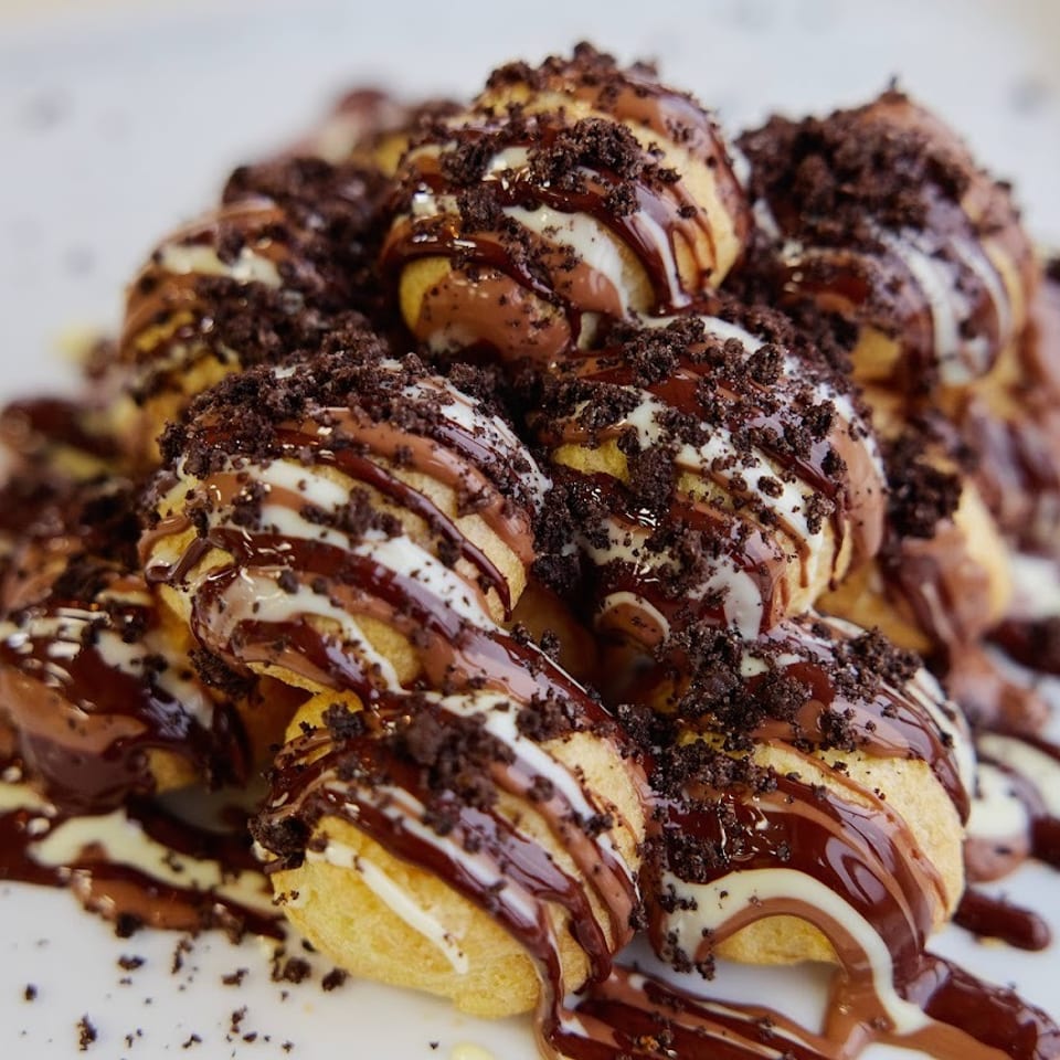 Golden pastry bites drizzled with chocolate and cream sauces, topped with crushed chocolate cookies on a white plate.
